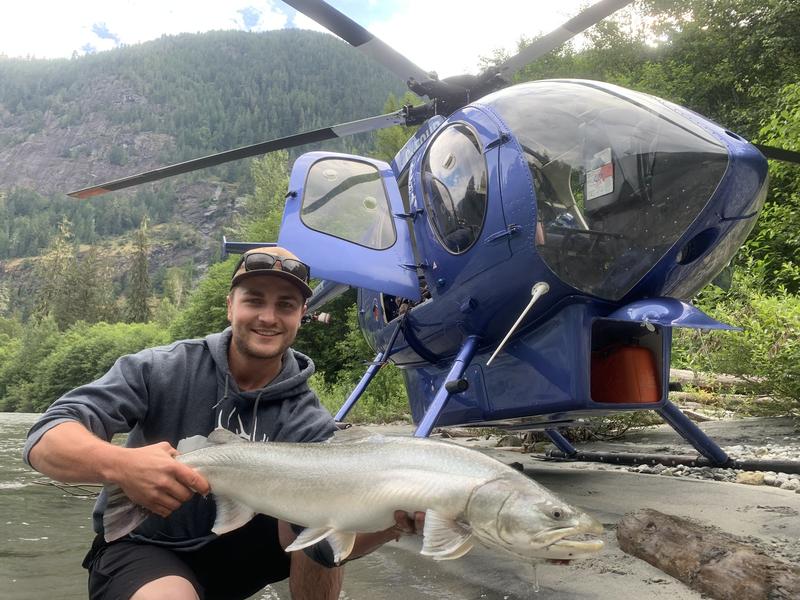 A man holding a large silver salmon in front of the open door of a blue helicopter.
