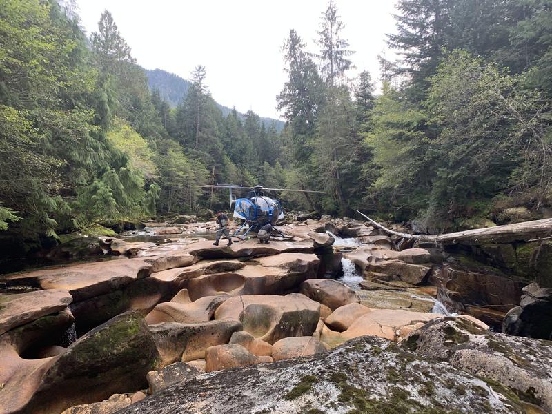A scenic view of a rocky riverbed with a blue helicopter landed in the distance near a forest.