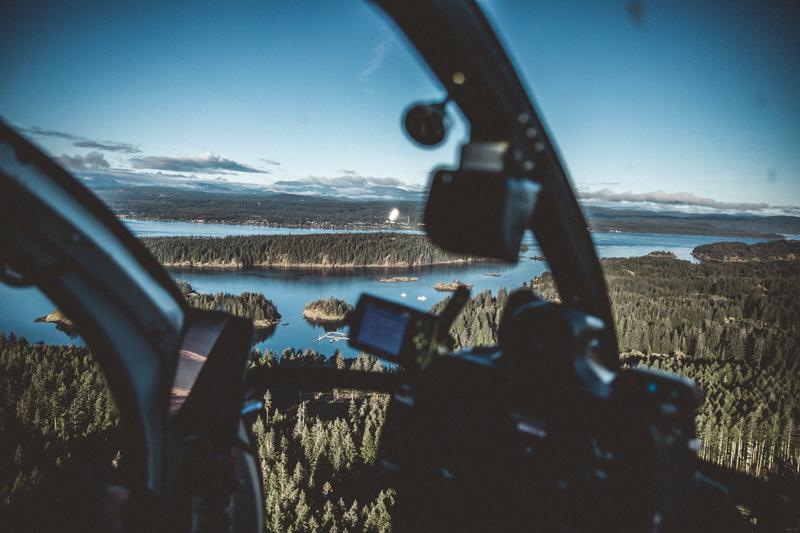 View from inside a helicopter cockpit looking out over a forested coastline and islands.