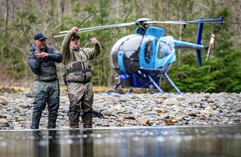 Two anglers standing in a river with fishing rods, with a blue helicopter parked on the rocky bank behind them.
