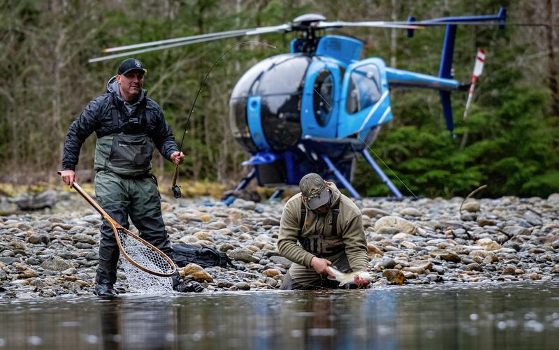 Two men on a riverbank near a blue helicopter; one stands with a net while the other crouches holding a fish.