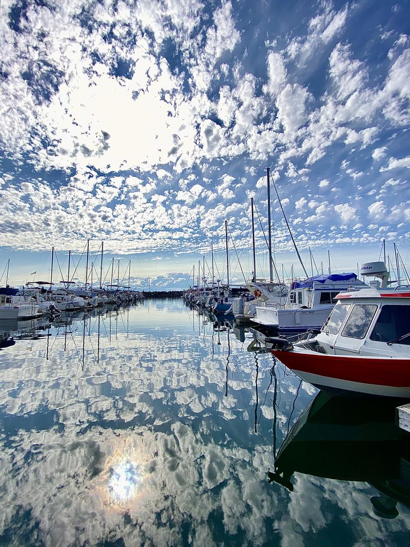 A marina filled with docked boats reflecting in calm water under a sky with scattered clouds.