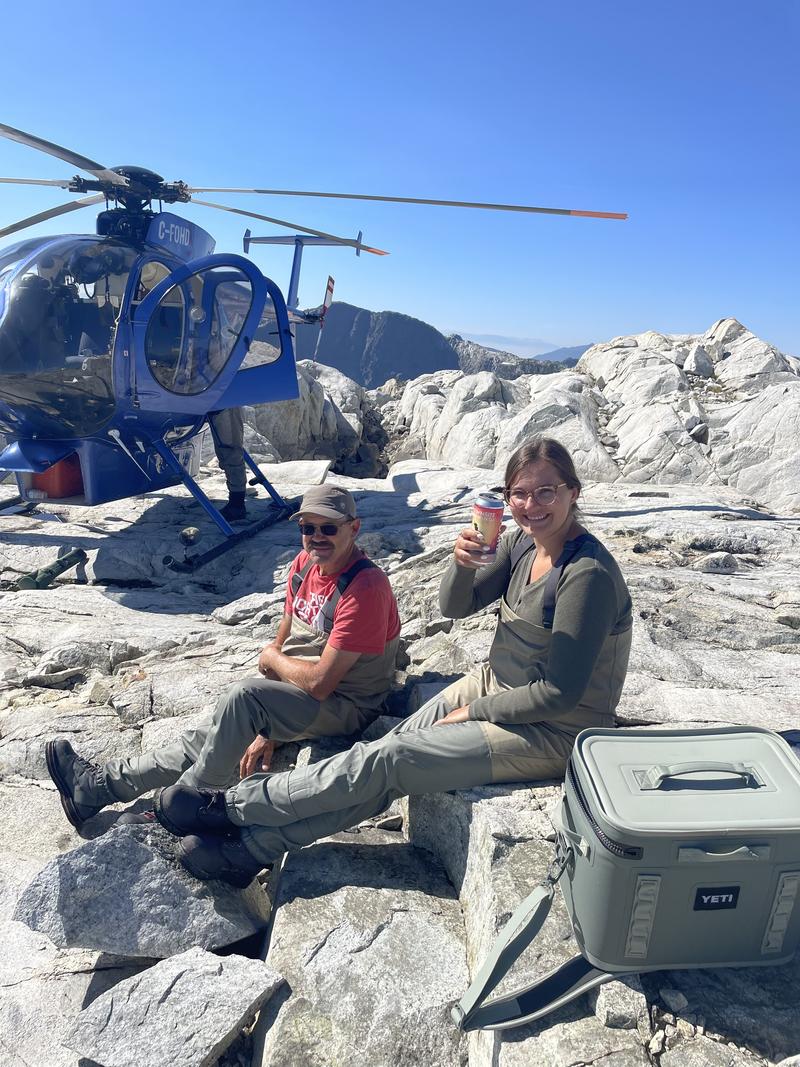 A man and a woman sitting on rocks near a blue helicopter, relaxing with drinks.