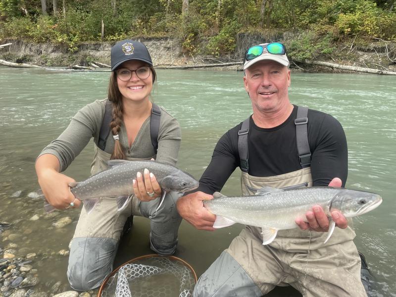 A woman and a man standing in a river, each holding a large salmon.