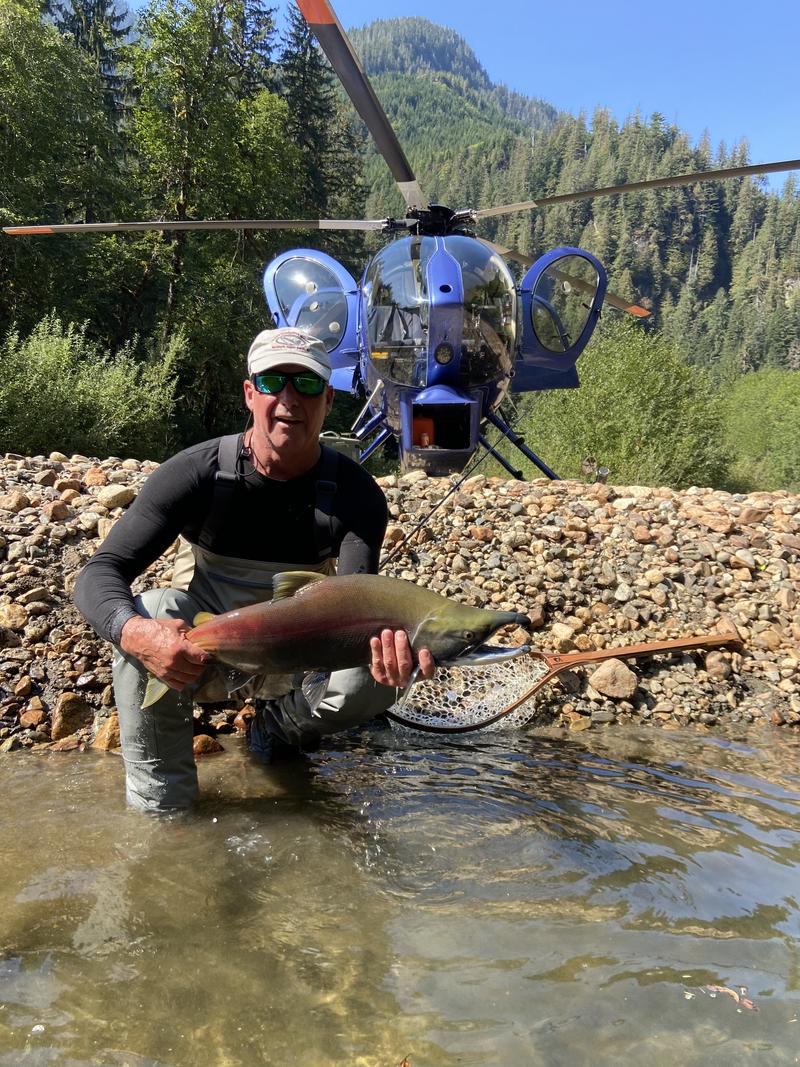 A man kneeling in a shallow river holding a fish, with a blue helicopter and rocky shore in the background.
