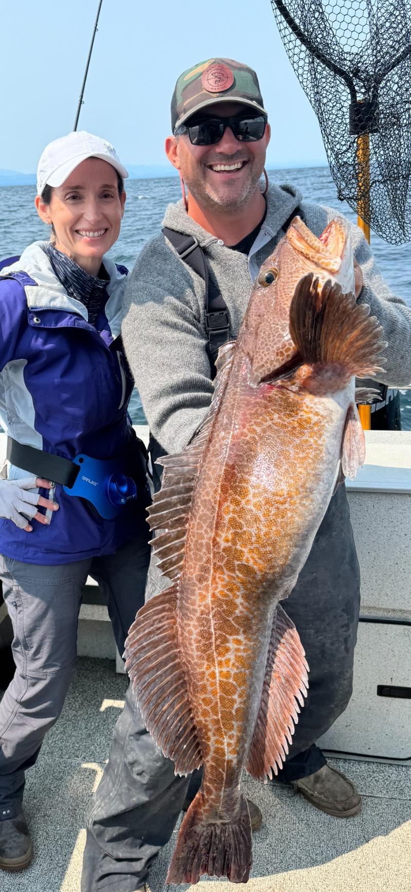A man and a woman on a boat holding a large lingcod together.