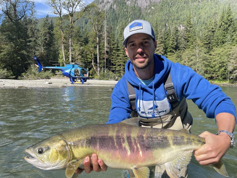 A man in a blue hoodie holding a large trout or char in a river, with a blue helicopter in the background.