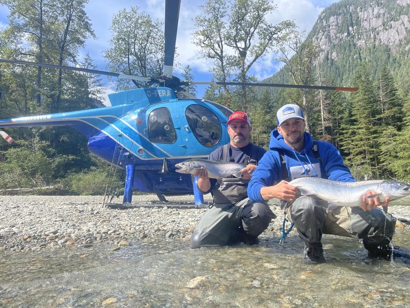 Two men kneeling in front of a blue helicopter, each holding a large salmon.