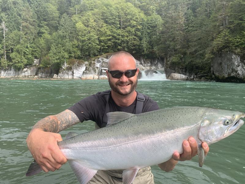 A smiling man in sunglasses holding a large silver salmon while standing in a river.